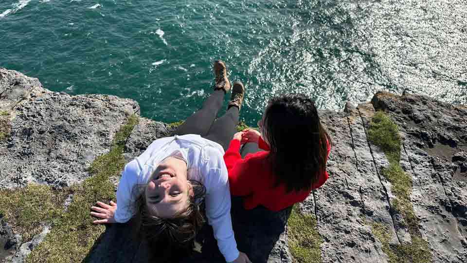 Lina sitzt am Rand einer Klippe und schaut kopfüber lachend in die Kamera